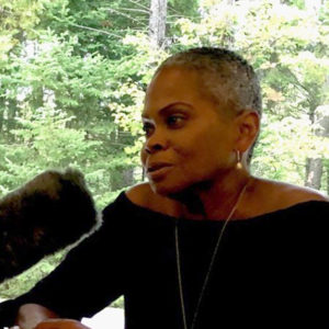 A photo of Bina Aspen-Rothblatt, who is wearing a black shirt and necklace and has a microphone pointing toward her.