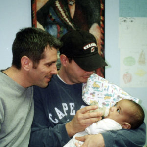 Two men holding a baby, gazing down at him and feeding him a bottle.