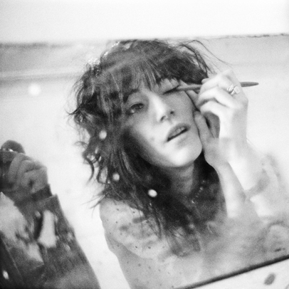 A black and white photograph of Patti Smith applying eye makeup with a pencil, all reflected in a mirror. In the corner of the photograph you can see a reflection of the photographer's arm holding the camera.