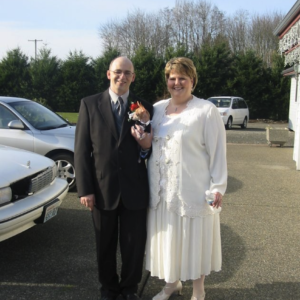 Jeff Ingram and Penny Hansen on their wedding day, standing together in a parking lot and smiling. Jeff is wearing a suit and holding a small dog, and Penny is wearing a white dress and cardigan.