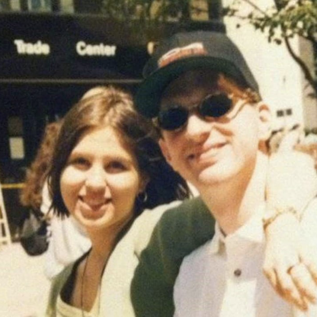 A young white couple posing outside on a sunny day. The man is wearing sunglasses and a hat. The woman has her arm around the man's shoulders and they're both smiling.