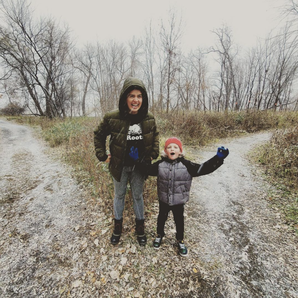 Kate Bowler, and her young son, Zach, standing at the fork of a trail outdoors in the snow. They’re bundled up wearing jackets and gloves, both smiling, and Zach has his arms spread wide open.