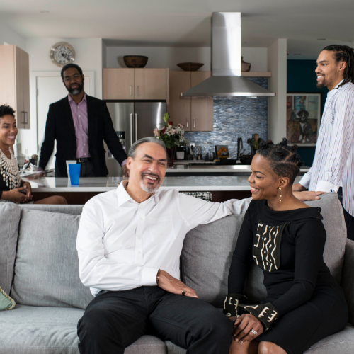 Phil and Nnenna Freelon sitting on a gray couch. Nnenna is looking at Phil. They both are smiling. Their three children Deen, Maya and Pierce are in the background.