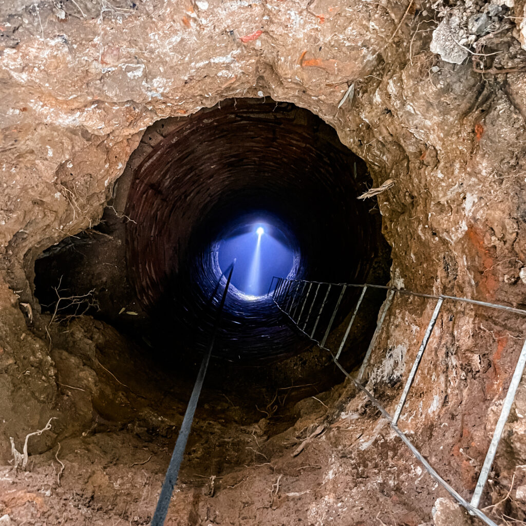 Photograph of someone standing at the bottom of a deep hole, wearing a headlamp and looking up and the camera. There's a metal ladder on the right hand side of the photo.