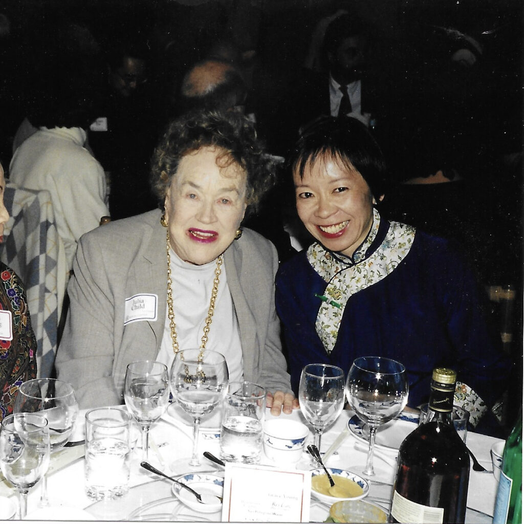 Photograph of two women sitting at a banquet dining table. There are wine glasses and plates on the table in front of them. The woman on the left is wearing a gray sweater. The woman on the right is wearing a black shirt and a scarf