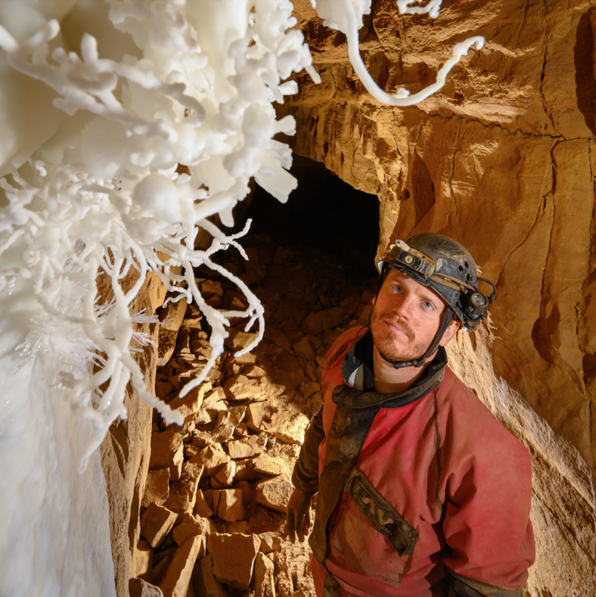 A man wearing a red jumpsuit and helmet stands in a cave. There is a large passageway behind him.