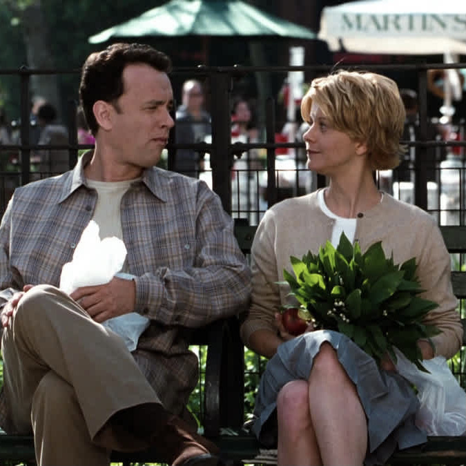 Still from the movie You’ve Got Mail, with Tom Hanks and Meg Ryan sitting on a park bench, looking at each other. He’s holding a plastic bag in his lap and she’s holding what looks like a plant.