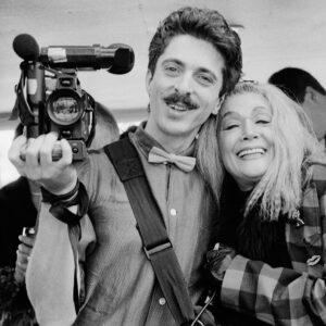 A black and white photograph of Nelson Sullivan holding a camera in one hand. His other hand is wrapped around Sylvia Miles. Nelson has a bow tie, Sylvia is wearing a checkered shirt. They're both smiling.