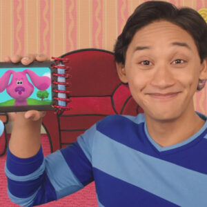 Color still of Josh Dela Cruz on the TV Show, Blue's Clues. He's wearing a striped blue shirt and holding a notebook with a picture of a cartoon dog.