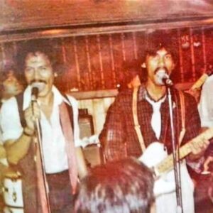 In an old photo, two young Filipino men stand in front of microphones, singing. Spanky Rigor is on the left, grinning at the audience while he sings. His bandmate is also singing while playing a guitar. The men have shoulder length hair and mustaches and wear button down shirts.