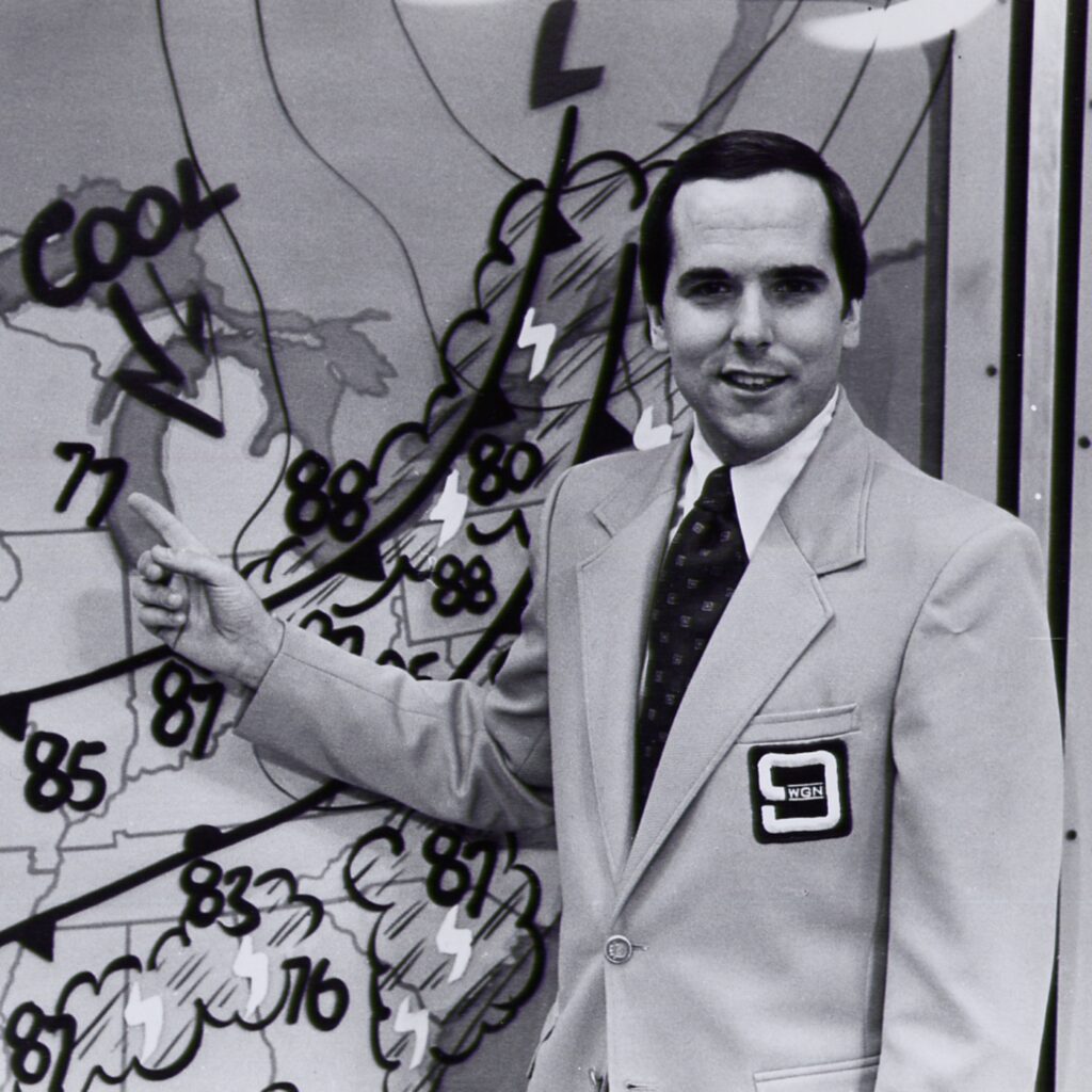 A black and white old photo of TV meteorologist Tom Skilling gesturing to a weather map behind him, with weather fronts and the word "cool" drawn on it. He is wearing a light colored suit with "WGN" and a "9" on a patch.