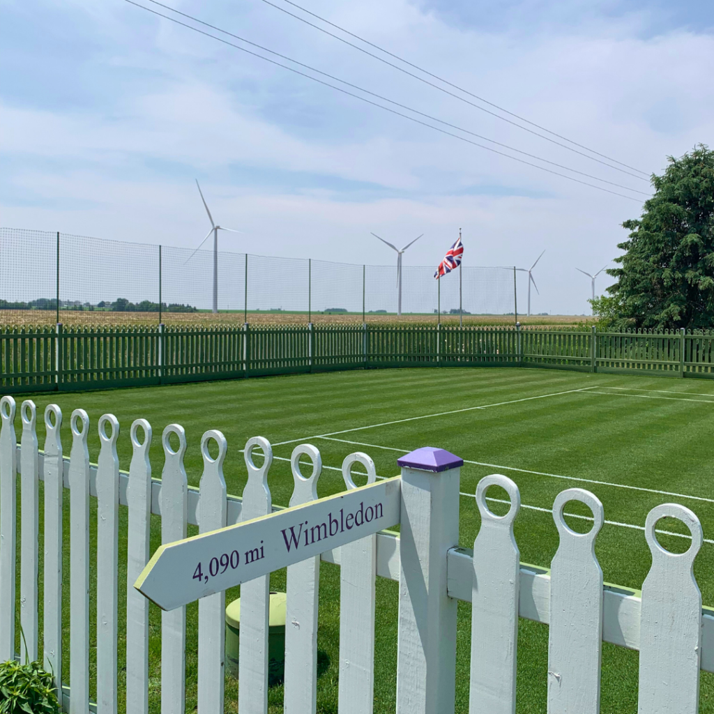 A grass tennis court surrounded by a white and green picket fence. In the foreground is a sign that reads “4,090 miles, Wimbledon.” Behind the tennis court is a British flag, tall white wind turbines, and corn fields.
