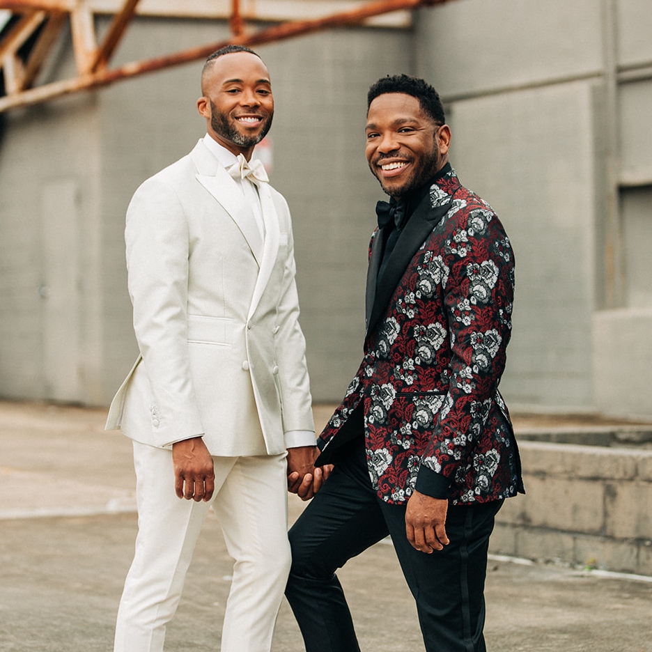 A photo of two men smiling while looking at the camera. They are standing, slightly facing each other, holding hands. One man is wearing a white suit and the other man is wearing black pants and a red, white and black floral suit jacket. Both men are wearing bowties and have facial hair.