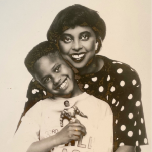 A black and white photo of Yla Eason behind her young son, Menelik Puryear. They are both smiling and Yla has her arms around Menelik. Menelik is holding a Sun-Man action figure.