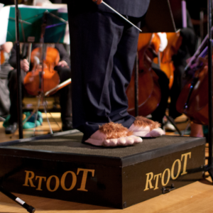 A photo of a conductor's legs standing on a podium. The conductor is wearing a suit and holding a baton, and on his feet are hobbit slippers - hairy feet with toes. The podium has the letters "RTOOT" on the sides.