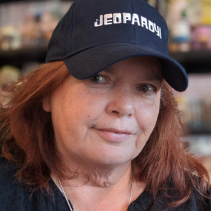 Jean Westcott wears a “Jeopardy!” hat and holds up a pen. Jean has long brown hair underneath a dark baseball cap. The baseball cap features the “Jeopardy” logo in white letters.