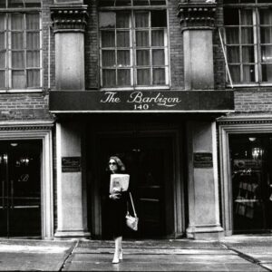 Black and white photo of the front of an apartment building with the words Barbizon Hotel on the awning. A woman carrying a stack of papers is walking away from the front door.