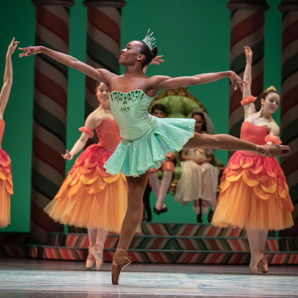 Ashton Edwards dancing the role of Dewdrop in "The Nutcracker," wearing a seafoam green short tutu with rhinestones and a sparkly tiara. Dancers in the background wear long orange and pink tutus.