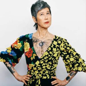 Valorie Lee Schaefer, an Asian woman with short black and white hair, standing against a white background with her hands on her hips, and wearing a blouse with contrasting flower patterns and a tattoo partly visible across her chest.