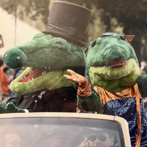 Kourtney and Brian LaPlant dressed up as Albert and Alberta, the University of Florida mascots, standing in the backseat of a car. It looks like they're in some kind of parade. Albert and Alberta are fuzzy green alligator costumes -- Albert is wearing a black suit and top hat and Alberta has a dress with blue sequins and orange gloves.