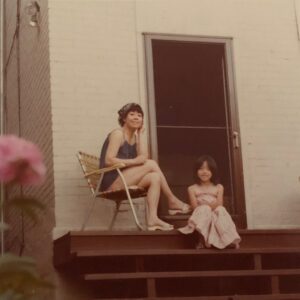 Kyo Maclear as a young girl sitting at the top of a set of brown stairs in front of a screen door looking over her backyard. Her mother, a Japanese woman wearing a bandana over short hair, is sitting in a deck chair next to Kyo. Both are wearing summer dresses. The photo is taken just behind a bush with pink flowers that are on the left edge of the photo.