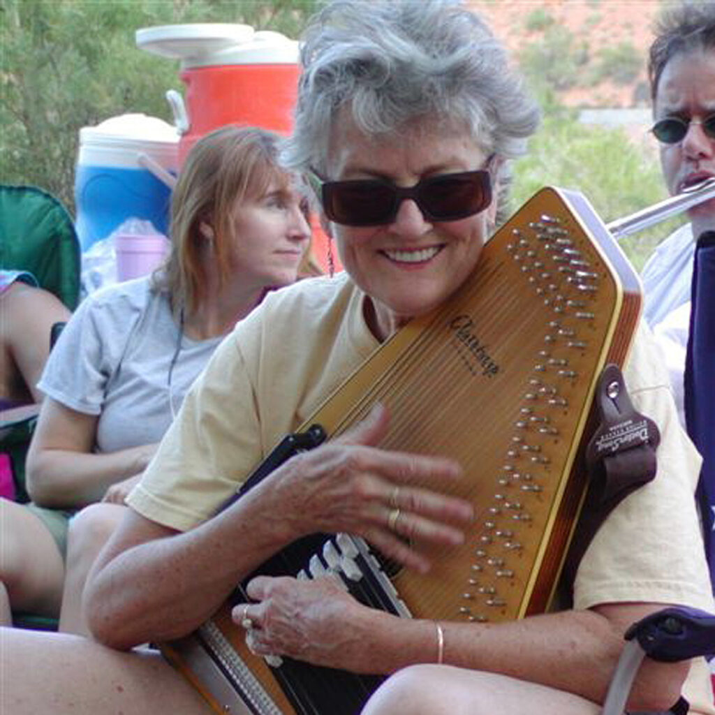 Color photograph of Peggy Seeger, a white woman with short light gray hair, sitting in a camp chair outdoors, playing an autoharp. She is wearing sunglasses and grinning at the camera. There are colorful water coolers and other people, including one playing a flute, behind her.