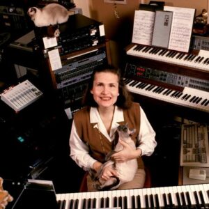 Photo of Wendy Carlos surrounded by electronic keyboards and synthesizers, with a cat sitting behind her and another cat in her lap.