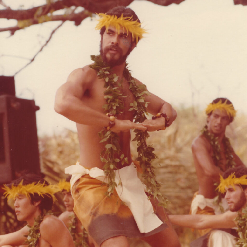 A sepia-toned photograph of a young Hawaiian man, Patrick Makuakāne, mid-dance. He is wearing a cloth skirt, a yellow lei around his head, and a lei of green leaves draped around his neck. His knees are bent in a slight squat, and he looks over his right shoulder, away from the camera. There are other men in identical dress dancing and sitting in the background.