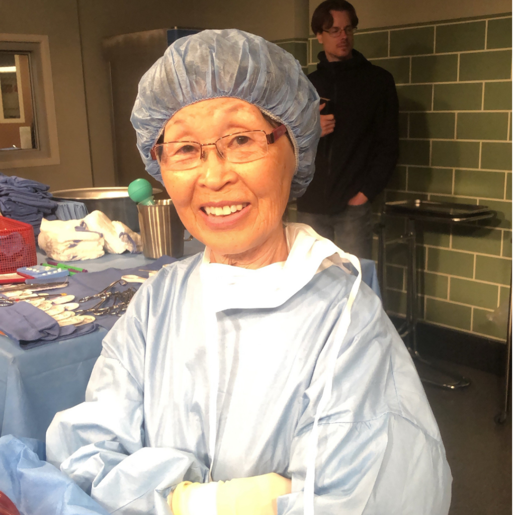 A photograph of BokHee An - an older Asian woman smiling and wearing glasses. She is standing in what looks like an operating room, wearing light blue scrubs, surgical gloves, a surgical hair cap and a surgical mask partially tied around her neck. There is a tray of surgical instruments laid out on a table behind her.