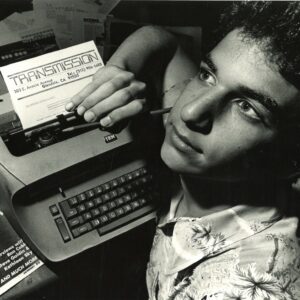 Black and white photo of Miguel Esteban as a teenager at his desk leaning over a typewriter with a piece of paper with the logo of his magazine, Transmission.