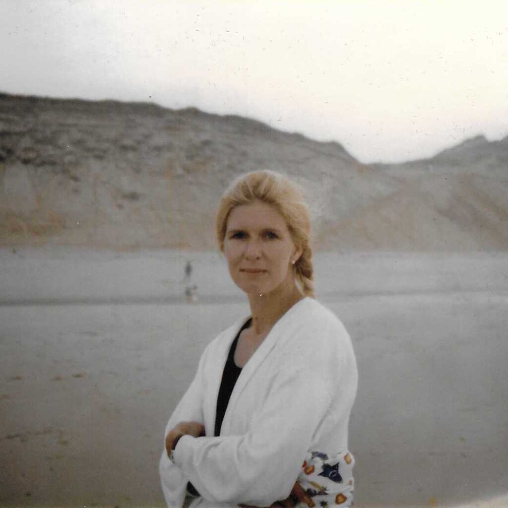 Photograph of Valentine Judge in her late 30s with a beach in the background. She's wearing a cardigan, has her hair in a braid, and is looking directly at the camera with a slight smile.