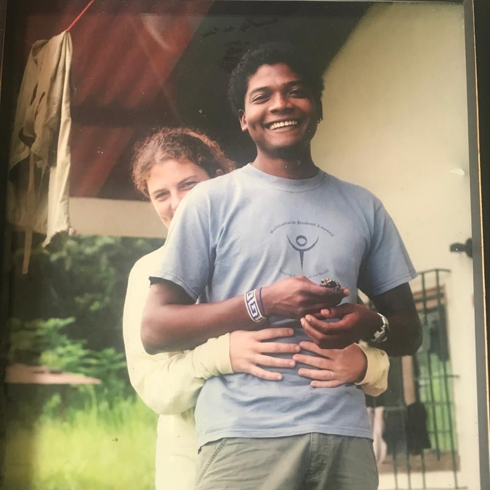 An old color photograph of a couple standing on a porch under a clothesline. In front, a young Black man, Edgardo Griffth, smiles widely at the camera and holds a small frog in his hand. A young white woman, Heidi Ross, stands behind him with smiling eyes, arms wrapped around his waist.