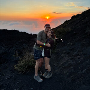 A photo of Haley Woloshen and Cody Bryant standing on a mountain in front of an orange sunset. They are both smiling and have their arms around each other.