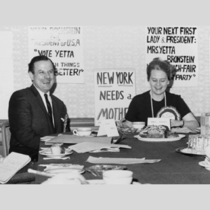 A black and white photo of two people sitting at a table laughing. One is a man dressed in a suit, and the other person is a woman. On the wall behind them are posters for "Your next first lady president, Mrs. Yetta Bronstein." They appear to be working: there are papers scattered around the table as well as a coffee cup and some cake.