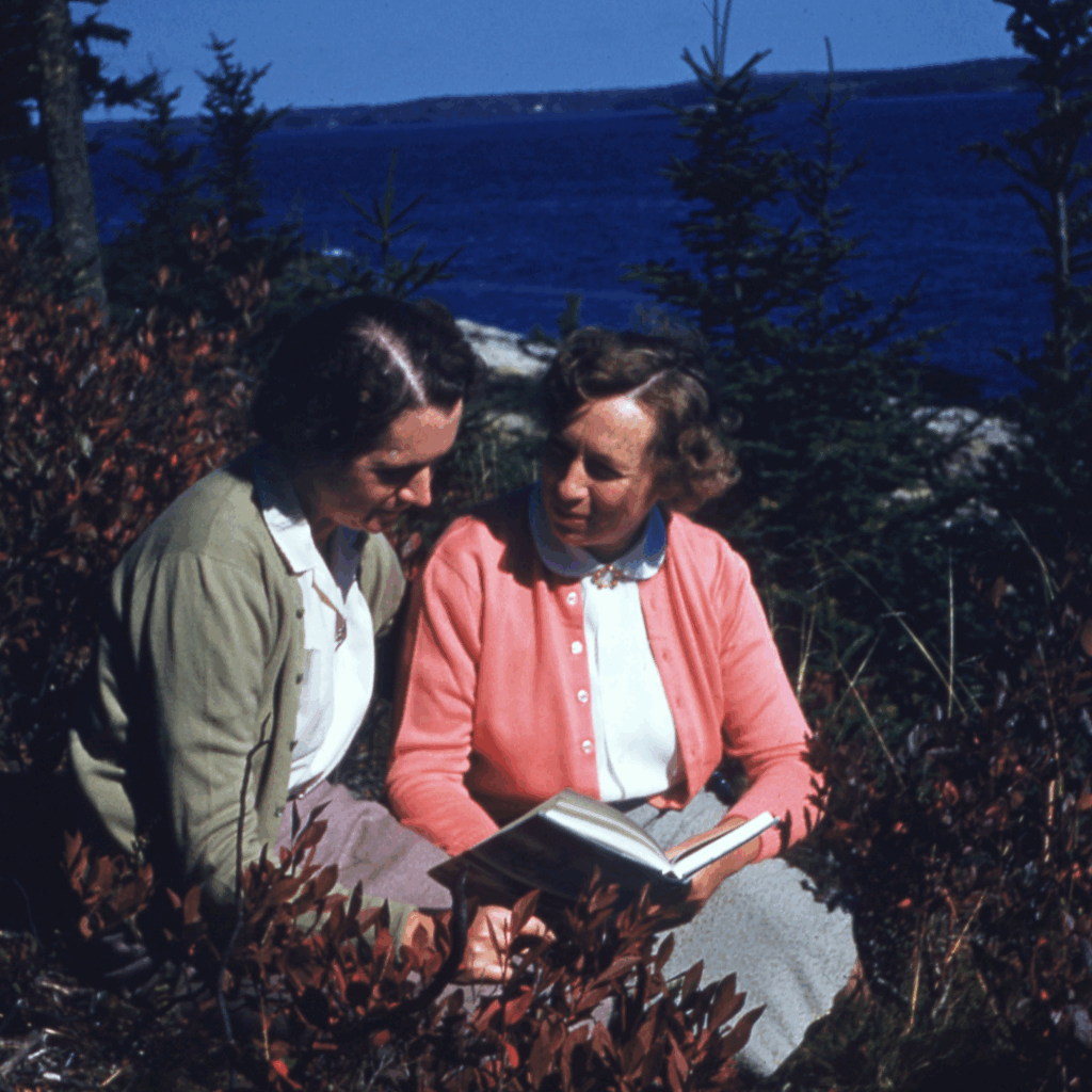 An old color photograph of Rachel Carson and Dorothy Freeman reading a book together. They're surrounded by trees and behind them is a blue ski.