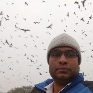 A selfie of Nadeem Shehzad. He's wearing glasses, a jacket, and a gray beanie, with a neutral expression. The sky behind him is gray and clouded and full of flying birds - black kites.