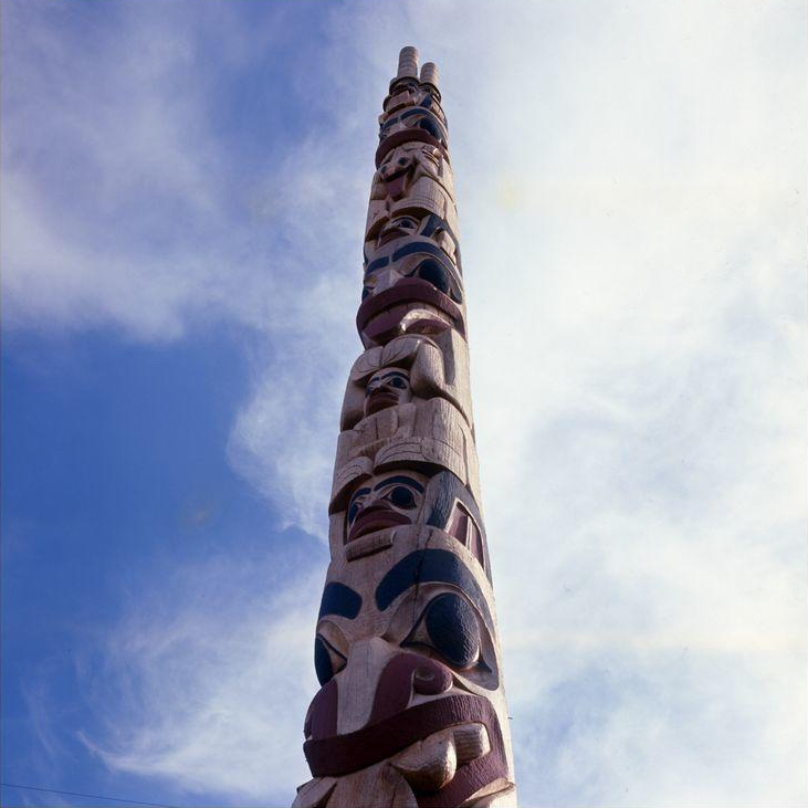 Color photograph of Robert Davidson's "Bear Mother" totem pole, taken from the ground looking up. The totem pole reaches into the sky and is covered in figures and faces painted in red and black paint. There are wispy clouds in the sky behind the totem pole.