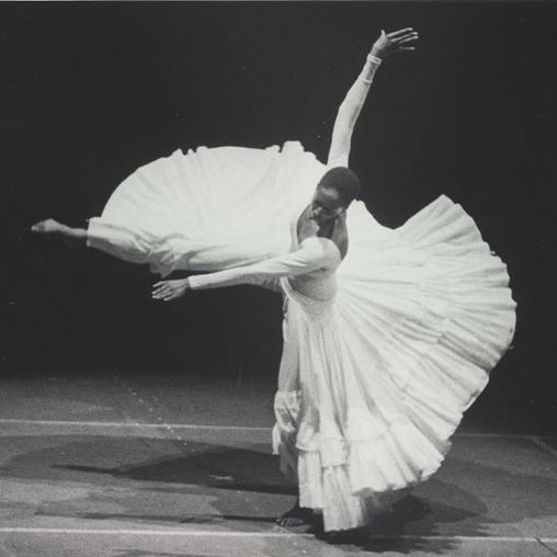 A black and white photograph of Judith Jamison, a Black woman, dancing “Cry.” She stands on her left leg, her right leg extended in the air to form an angle about ninety degrees wide, toe pointed. Her torso is twisted, her head angled partially toward the floor. Her right arm extends vertically above her head and her left arm forms a straight line just slightly below her right leg. Her full ankle-length white skirt arcs around her, forming an approximate three-quarters circle. She is barefoot and wearing a white long-sleeve leotard with her skirt. Her hair is short. The stage behind her fades to black. Photo credit: Fred Fehl, Jerome Robbins Dance Division, The New York Public Library.
