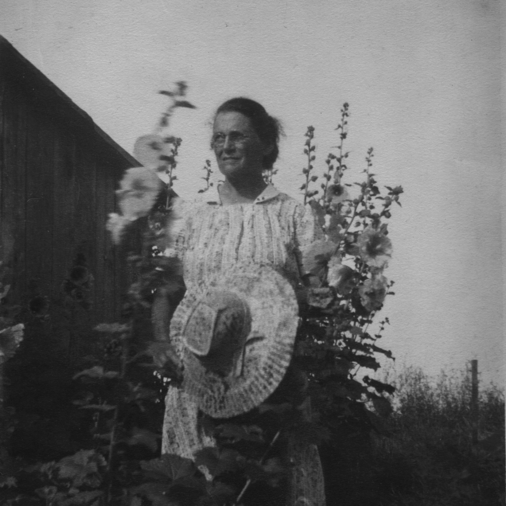 Black and white photo of Emma Gatewood at age 54. There are flowers in the foreground, framing her, as if Emma is standing in a garden. She is wearing a dress and holding a wide brimmed hat. Her hair is pulled back in a bun and she looks off to her right, away from the camera. In the background there is a fence and an empty sky.