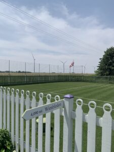 Color photograph of a grass tennis court in the middle of a cornfield with a United Kingdom flag in the background and a pointed sign that reads "4,090 mi Wimbledon"