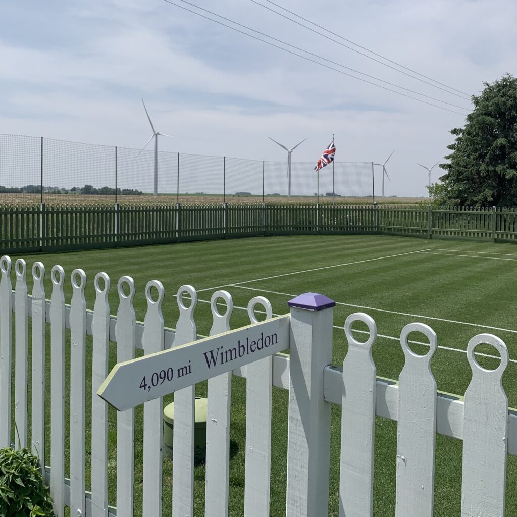 Color photograph of a grass tennis court in the middle of a cornfield with a United Kingdom flag in the background and a pointed sign that reads "4,090 mi Wimbledon"