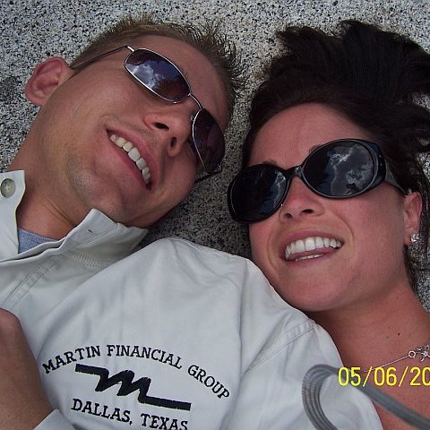 Close-up selfie of a young white couple laying down on a speckled rocky surface. They are both wearing sunglasses which reflect the clouds and they are smiling, slightly flushed. The woman has long brown hair and the man has short blonde hair. A logo on his shirt reads “Martin Financial Group” and “Dallas, Texas.” A yellow date stamp in the corner reads 05/06/2006.