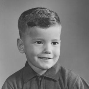 An old black and white photograph of a young boy around the age of 5 with short, combed hair. He is wearing a polo shirt, smiling at the camera.