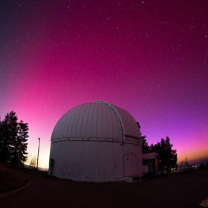 An observatory building at the top of Mount Lemmon outside of Tucson, Arizona. The observatory is a small white circular building with a domed roof. The evening sky behind it is dark pink and purple, and you can see hundreds of stars.