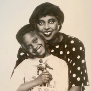 Black and white photograph of Yla Eason and her young son Menelik. Yla has her arms around Menelik, who is holding the Sun-Man action hero toy. Both are smiling. Yla is a Black woman with short hair wearing a polka dot shirt, and Menelik is wearing a t-shirt.