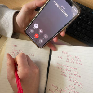 Color photograph of someone holding an iPhone in one hand while on a call, and taking notes in a notebook with the other hand. Some of the visible notes read: New entanglement, thick green line.
