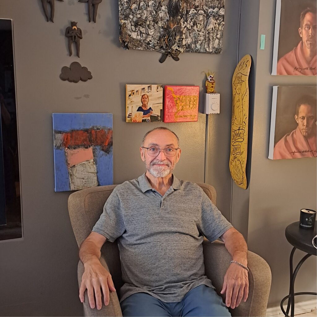 Bill Staubi sitting in an armchair in his apartment. He is wearing a gray polo shirt and glasses. He smiles at the camera. On the wall behind him hangs various types of art works: paintings, small sculptures and a painted skateboard.